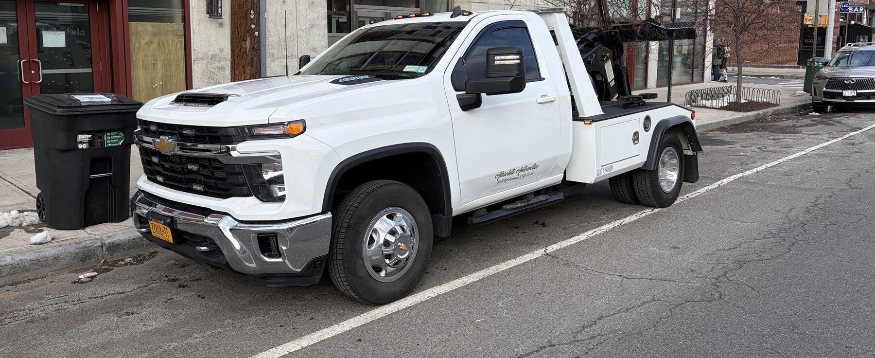 A white Absolute Automotive self-loading wrecker with gold company lettering on the door, parked curbside on a New York City street.